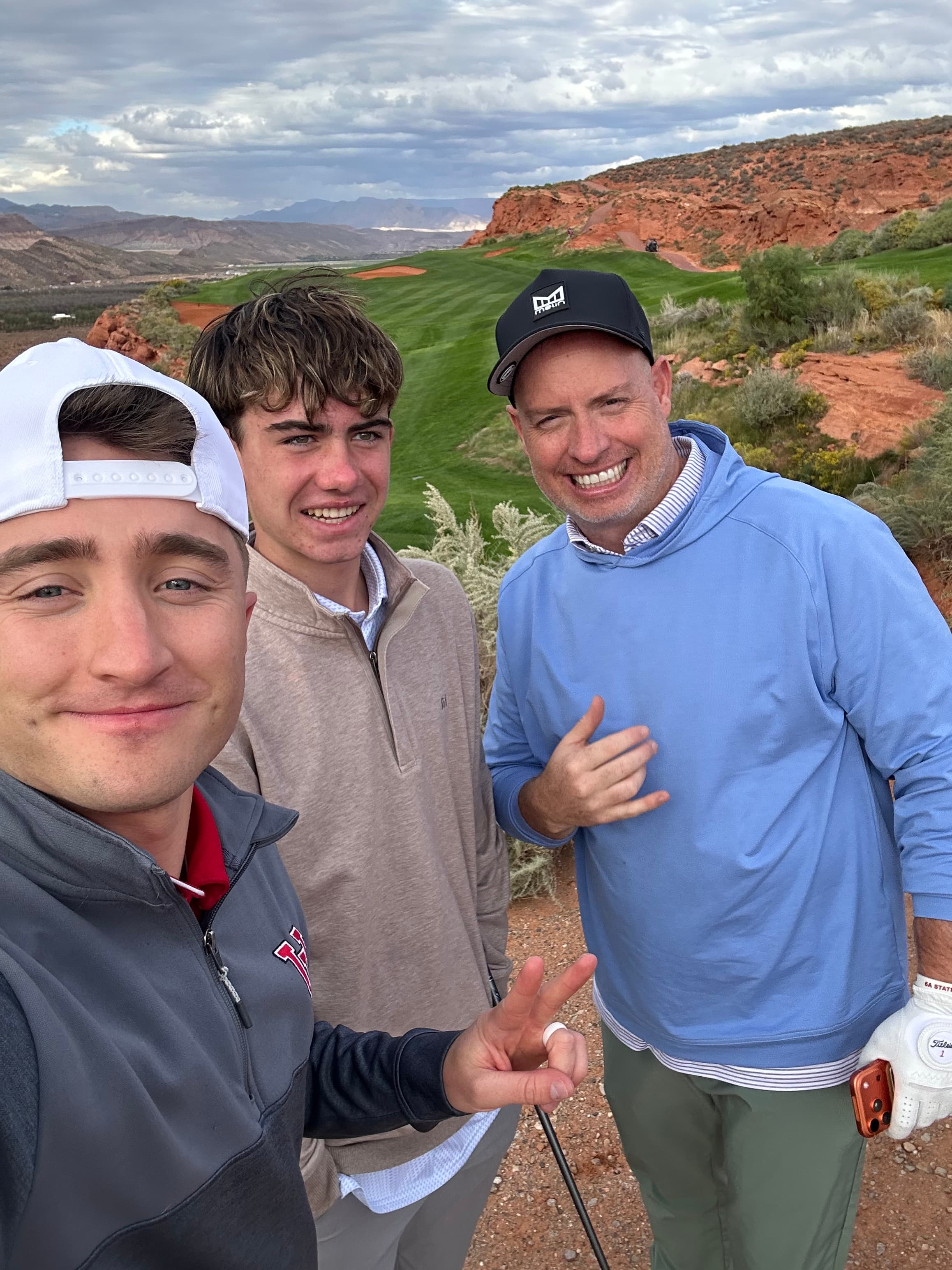 Three men smiling for a selfie on a golf course with scenic red rock cliffs.